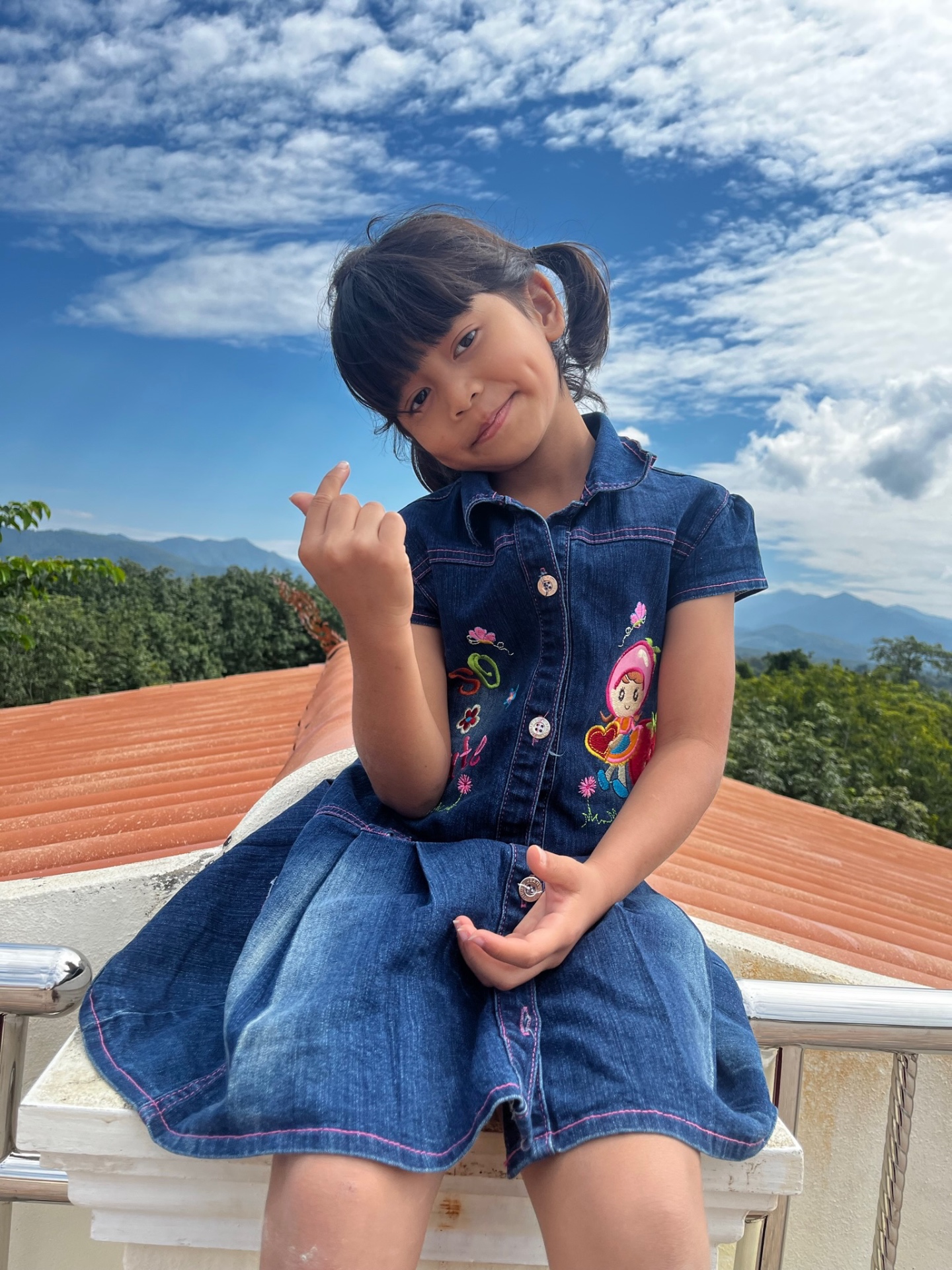 Girl in a denim dress on a rooftop with mountains and clouds behind her