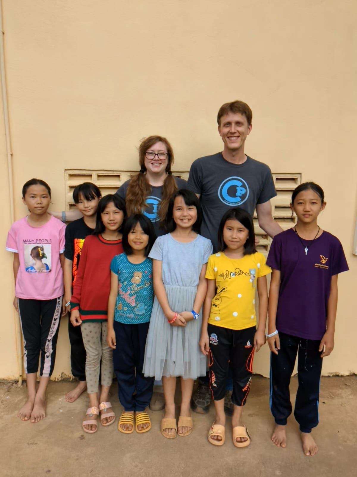 Two team members standing with a group of older Lanna girls against a yellow wall