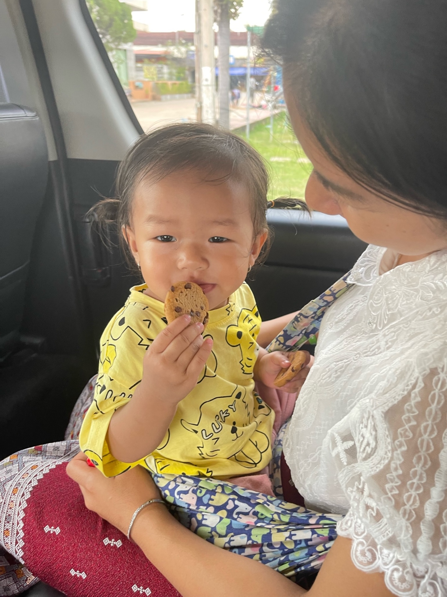 Baby in a yellow shirt holding a chocolate-chip cookie, on her mother's lap in a van