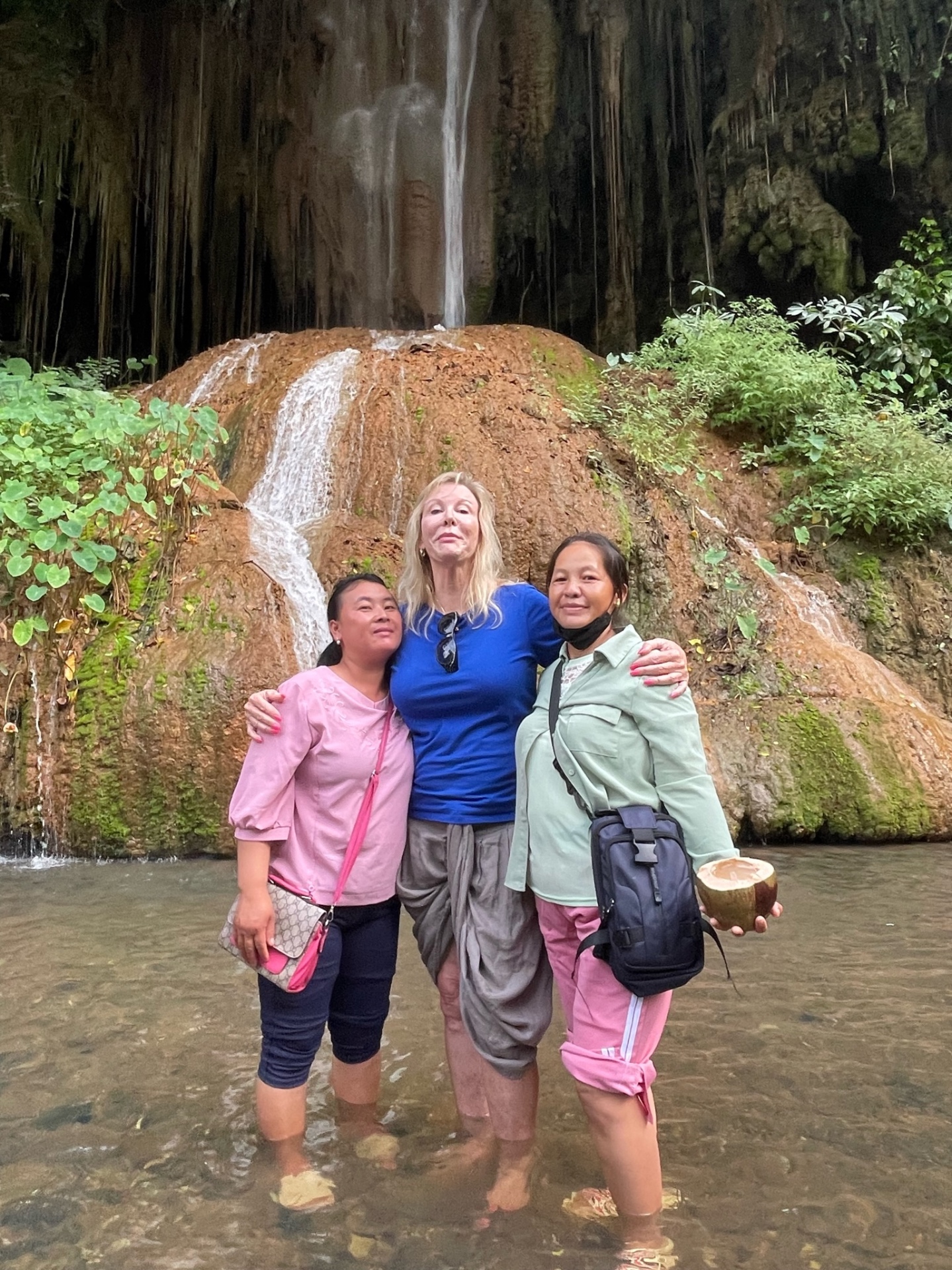 Three women — two Thai partners and a team member — standing in the stream beneath a waterfall